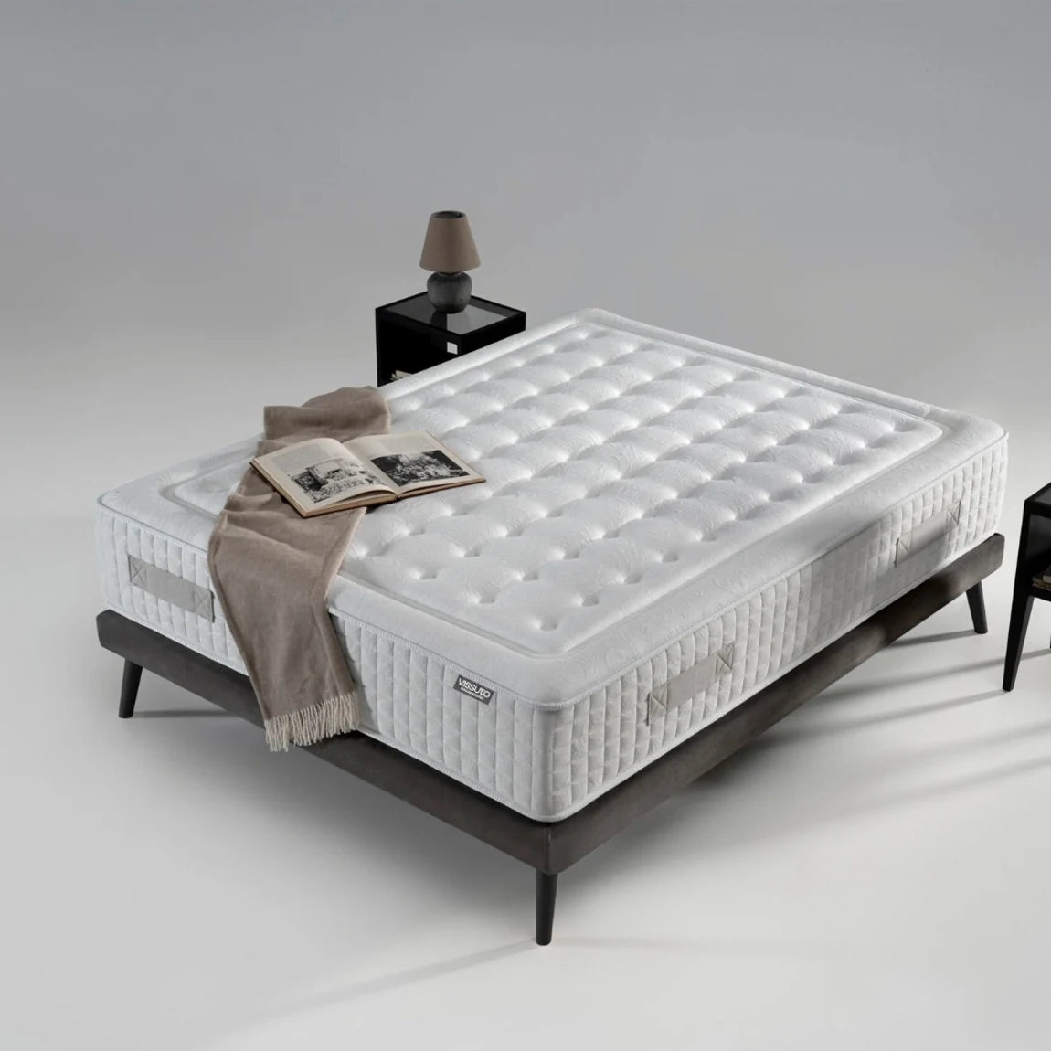 Colchón Vissuto Innova Pocket en cama con patas de madera oscura, mantas y mesita de noche en dormitorio minimalista